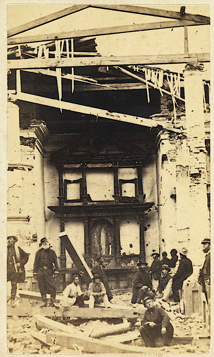 Fotografia antiga do interior de uma igreja destruída, com o telhado aberto, vigas penduradas, um altar ao fundo e um grupo de soldados de pé entre os escombros