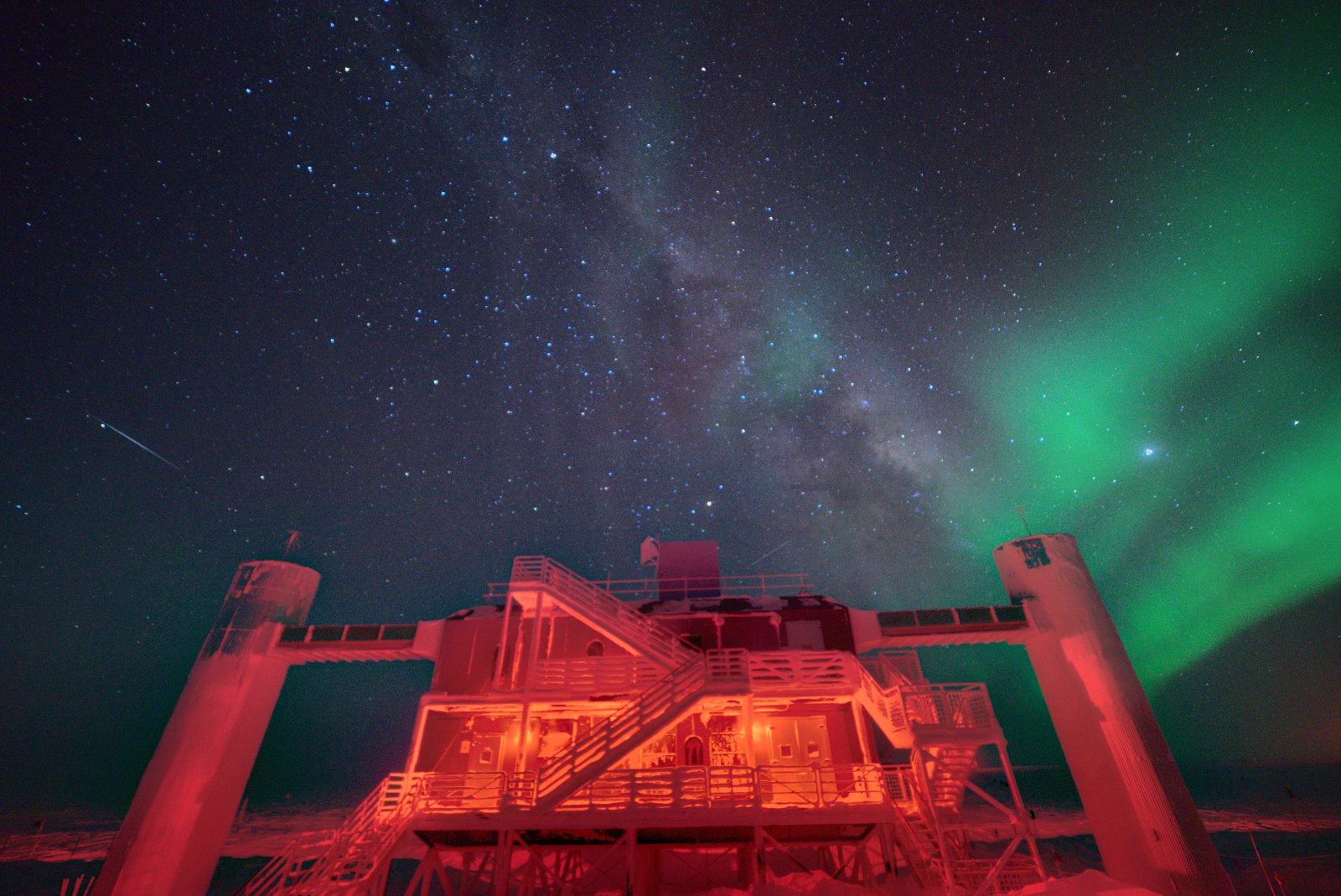 Prédio do laboratório IceCube sobre pilares, iluminado de vermelho, sob um céu noturno com a Via Láctea e uma aurora verde à direita