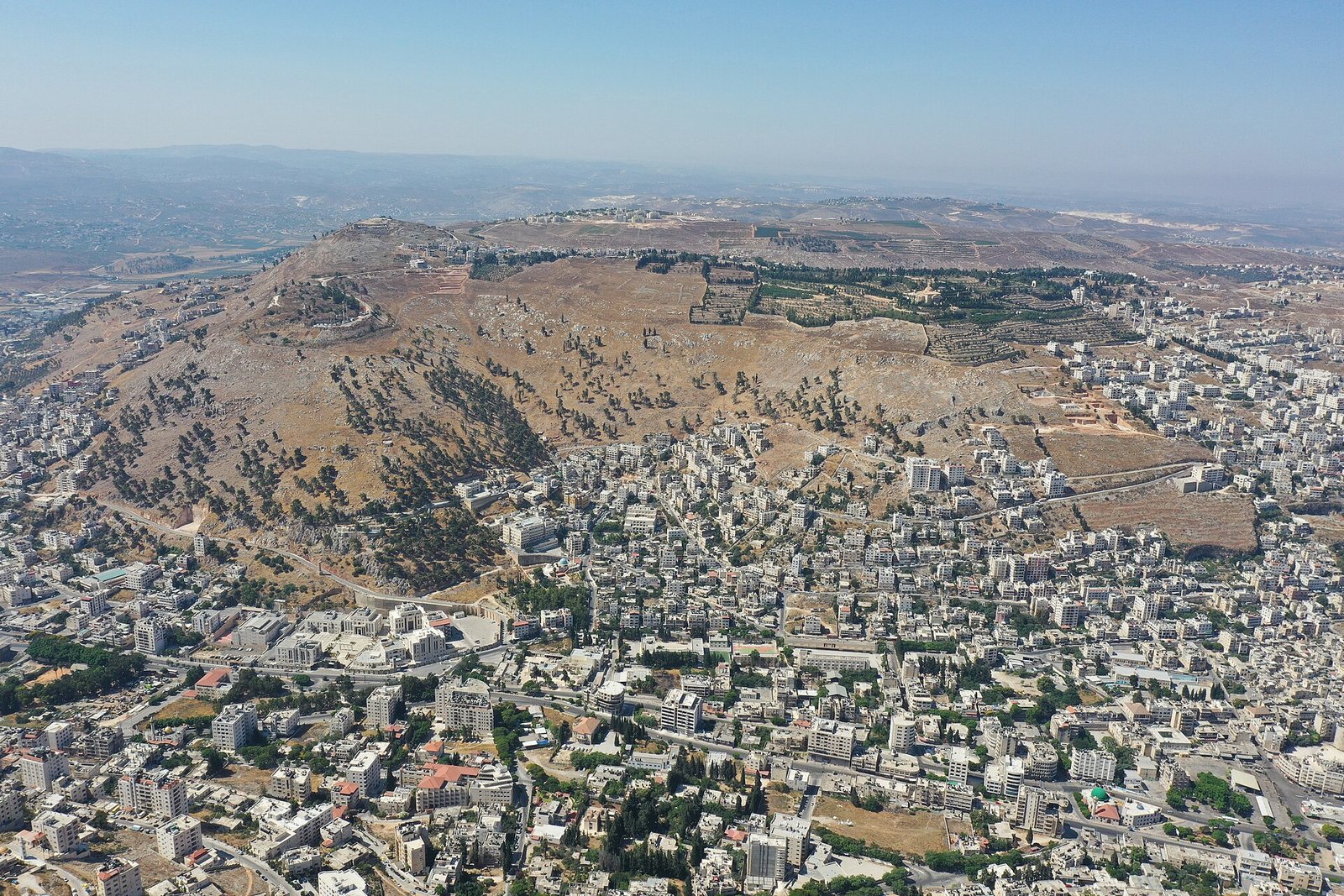 Vista aérea do monte Gerizim, coberto de vegetação rala, com a cidade de Nablus espalhada pelo vale aos pés da montanha