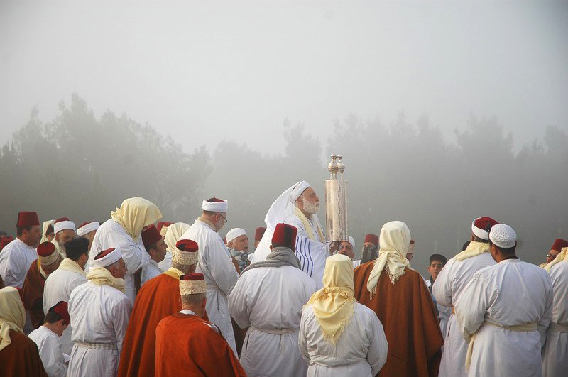 Grupo de homens samaritanos vestidos de branco, vários com barretes vermelhos, reunidos ao ar livre numa manhã de neblina, enquanto um deles ergue um objeto ritual